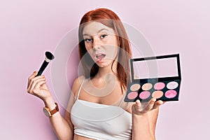 Young red head girl holding makeup brush and blush palette in shock face, looking skeptical and sarcastic, surprised with open