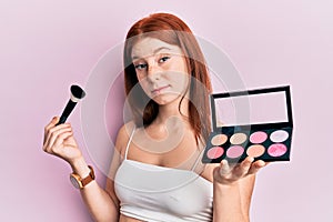 Young red head girl holding makeup brush and blush palette skeptic and nervous, frowning upset because of problem