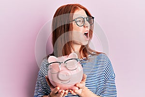 Young red head girl holding piggy bank with glasses angry and mad screaming frustrated and furious, shouting with anger