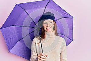 Young red head girl holding purple umbrella wearing fresh beret looking positive and happy standing and smiling with a confident