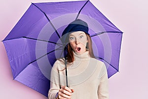 Young red head girl holding purple umbrella wearing fresh beret scared and amazed with open mouth for surprise, disbelief face