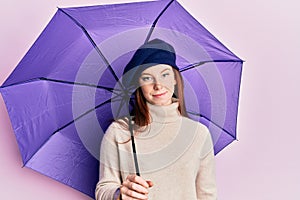 Young red head girl holding purple umbrella wearing fresh beret thinking attitude and sober expression looking self confident