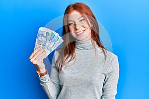 Young red head girl holding south african 100 rand banknotes looking positive and happy standing and smiling with a confident