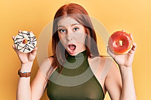 Young red head girl holding tasty colorful doughnuts afraid and shocked with surprise and amazed expression, fear and excited face