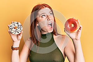 Young red head girl holding tasty colorful doughnuts angry and mad screaming frustrated and furious, shouting with anger looking