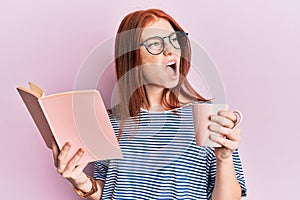 Young red head girl reading a book and drinking a cup of coffee angry and mad screaming frustrated and furious, shouting with