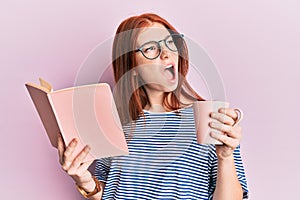 Young red head girl reading a book and drinking a cup of coffee angry and mad screaming frustrated and furious, shouting with