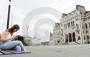 Young red head girl sitting in a bench at the parliament square at Budapest, Hungary. Teenager traveler holding a cellphone photo