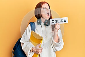 Young red head girl wearing backpack holding online banner angry and mad screaming frustrated and furious, shouting with anger