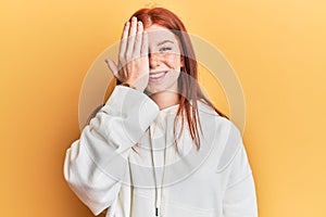 Young red head girl wearing casual sweatshirt covering one eye with hand, confident smile on face and surprise emotion