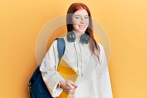 Young red head girl wearing student backpack and holding book looking positive and happy standing and smiling with a confident