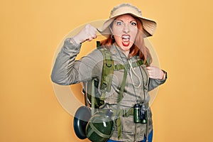 Young redhead backpacker woman hiking wearing backpack and hat over yellow background angry and mad raising fist frustrated and