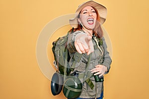 Young redhead backpacker woman hiking wearing backpack and hat over yellow background laughing at you, pointing finger to the