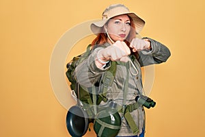 Young redhead backpacker woman hiking wearing backpack and hat over yellow background Punching fist to fight, aggressive and angry