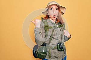 Young redhead backpacker woman hiking wearing backpack and hat over yellow background Surprised pointing with hand finger to the