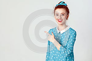 Young redhead caucasian girl with hair bun pointing her index finger away, indicating copy space on white blank wall for your cont