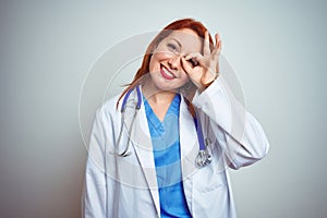 Young redhead doctor woman using stethoscope over white isolated background doing ok gesture with hand smiling, eye looking