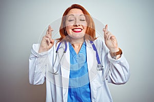 Young redhead doctor woman using stethoscope over white isolated background gesturing finger crossed smiling with hope and eyes