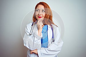 Young redhead doctor woman using stethoscope over white isolated background with hand on chin thinking about question, pensive