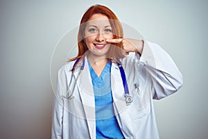 Young redhead doctor woman using stethoscope over white isolated background Pointing with hand finger to face and nose, smiling