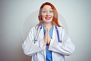 Young redhead doctor woman using stethoscope over white isolated background praying with hands together asking for forgiveness