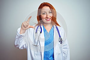 Young redhead doctor woman using stethoscope over white isolated background smiling and confident gesturing with hand doing small