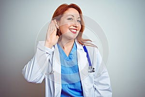 Young redhead doctor woman using stethoscope over white isolated background smiling with hand over ear listening an hearing to