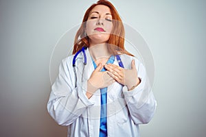 Young redhead doctor woman using stethoscope over white isolated background smiling with hands on chest with closed eyes and