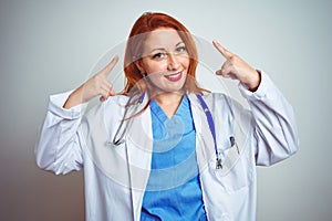 Young redhead doctor woman using stethoscope over white isolated background smiling pointing to head with both hands finger, great