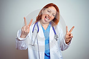 Young redhead doctor woman using stethoscope over white isolated background smiling with tongue out showing fingers of both hands