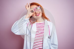 Young redhead doctor woman using stethoscope standing over isolated pink background doing ok gesture with hand smiling, eye
