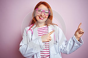 Young redhead doctor woman using stethoscope standing over isolated pink background smiling and looking at the camera pointing