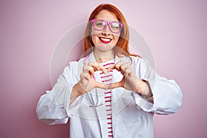 Young redhead doctor woman using stethoscope standing over isolated pink background smiling in love showing heart symbol and shape