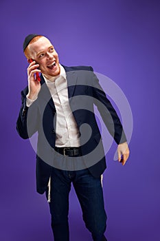 Young redhead emotional Jewish man emotionally talking on phone, greeting with holiday against purple studio background