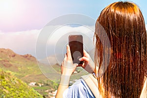 Young redhead girl photographer holding phone and taking a picture of landscape. Touristic concept. Mountains and blue