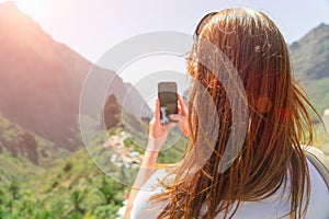 Young redhead girl photographer holding phone and taking a picture of Masca valley landscape. Sunny day over mountains