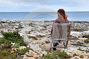 Young redhead girl sits on old wooden beach seat on the rocky sea coast