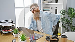 Young redhead guy with glasses suffering backache, anxiously touching injured spine while working indoors, sitting at the office