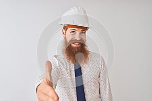 Young redhead irish architect man wearing security helmet over isolated white background smiling friendly offering handshake as