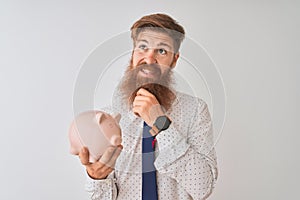 Young redhead irish businessman holding piggy bank over isolated white background serious face thinking about question, very