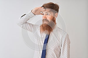 Young redhead irish businessman standing over isolated white background very happy and smiling looking far away with hand over