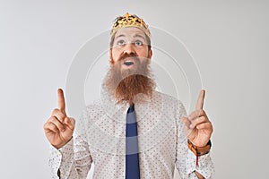 Young redhead irish businessman wearing crown king over isolated white background amazed and surprised looking up and pointing