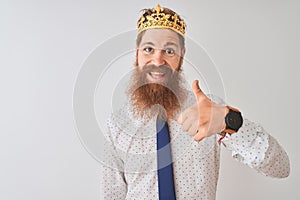 Young redhead irish businessman wearing crown king over isolated white background doing happy thumbs up gesture with hand