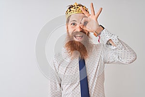 Young redhead irish businessman wearing crown king over isolated white background doing ok gesture with hand smiling, eye looking