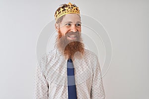 Young redhead irish businessman wearing crown king over isolated white background looking away to side with smile on face, natural