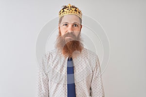 Young redhead irish businessman wearing crown king over isolated white background with serious expression on face