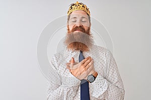 Young redhead irish businessman wearing crown king over isolated white background smiling with hands on chest with closed eyes and