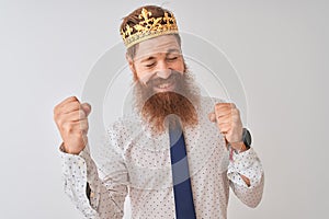 Young redhead irish businessman wearing crown king over isolated white background very happy and excited doing winner gesture with