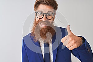 Young redhead irish businessman wearing suit and glasses over isolated white background doing happy thumbs up gesture with hand