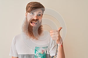 Young redhead irish man wearing t-shirt standing over isolated white background pointing and showing with thumb up to the side